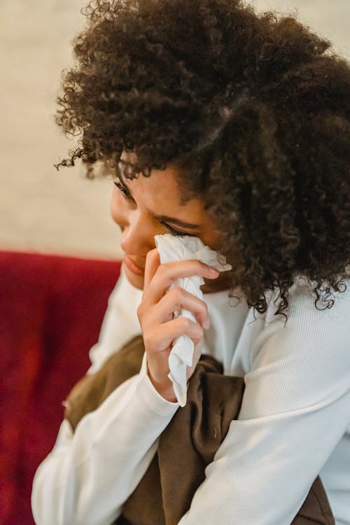 Depressed African American female with black curly hair wiping tears with tissue while sitting on sofa with cushion at home