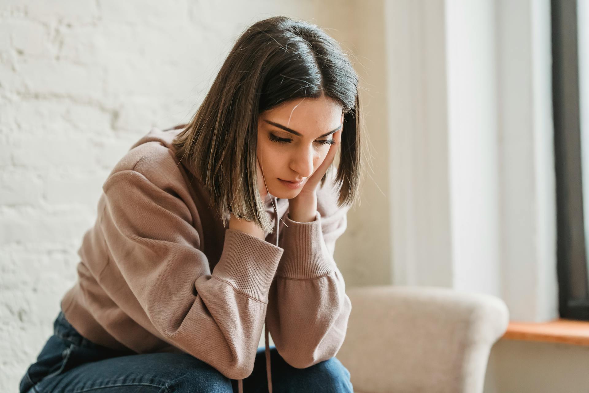 Unhappy female in casual wear touching face and looking down while sitting on sofa in light living room at home