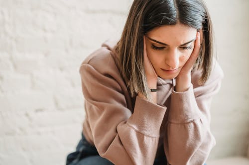 Free Crop unhappy female in casual wear touching cheeks and looking down while sitting near white wall in light room at home Stock Photo