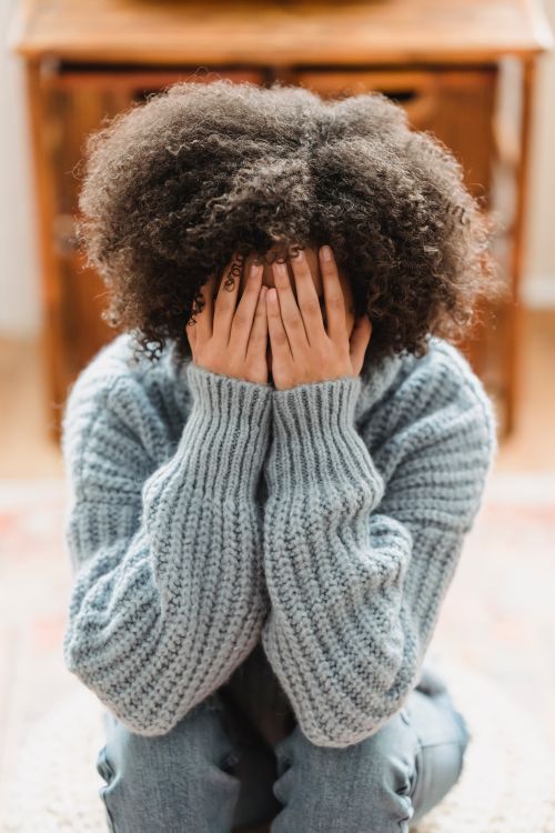 Free Unrecognizable sad African American female with curly hair hiding face with hands while sitting in light room at home on blurred background Stock Photo