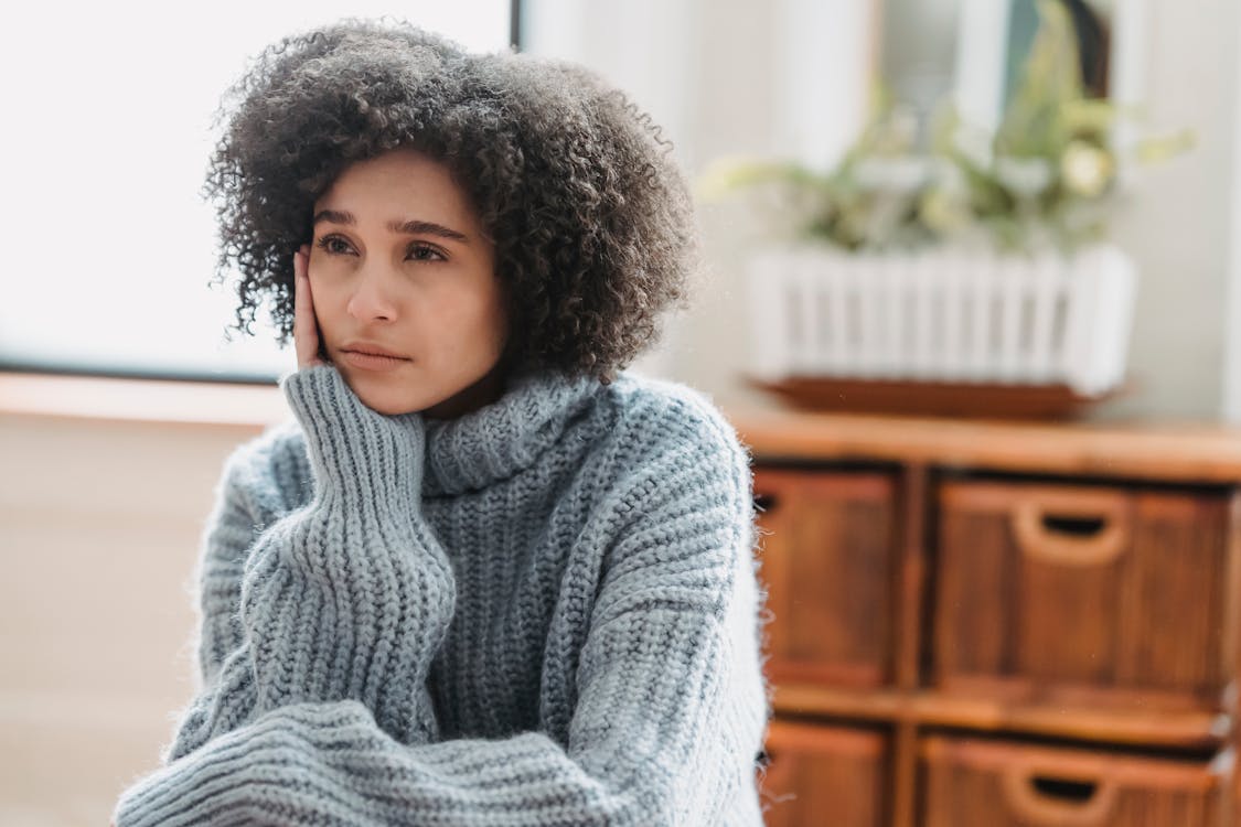 Free Discontent African American female leaning on hand and looking away while sitting in light room near green plant on blurred background Stock Photo