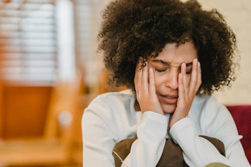 Despaired African American female with closed eyes touching face while sitting with pillow in light room at home on blurred background