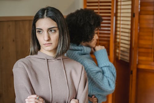 Free Upset multiracial women in casual wear standing in light room near wall with wooden folding screen while having conflict at home Stock Photo
