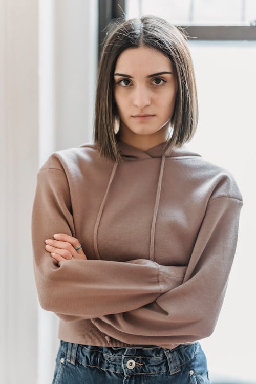 Dissatisfied female in casual wear looking at camera with serious face while standing with crossed arms in light room at home