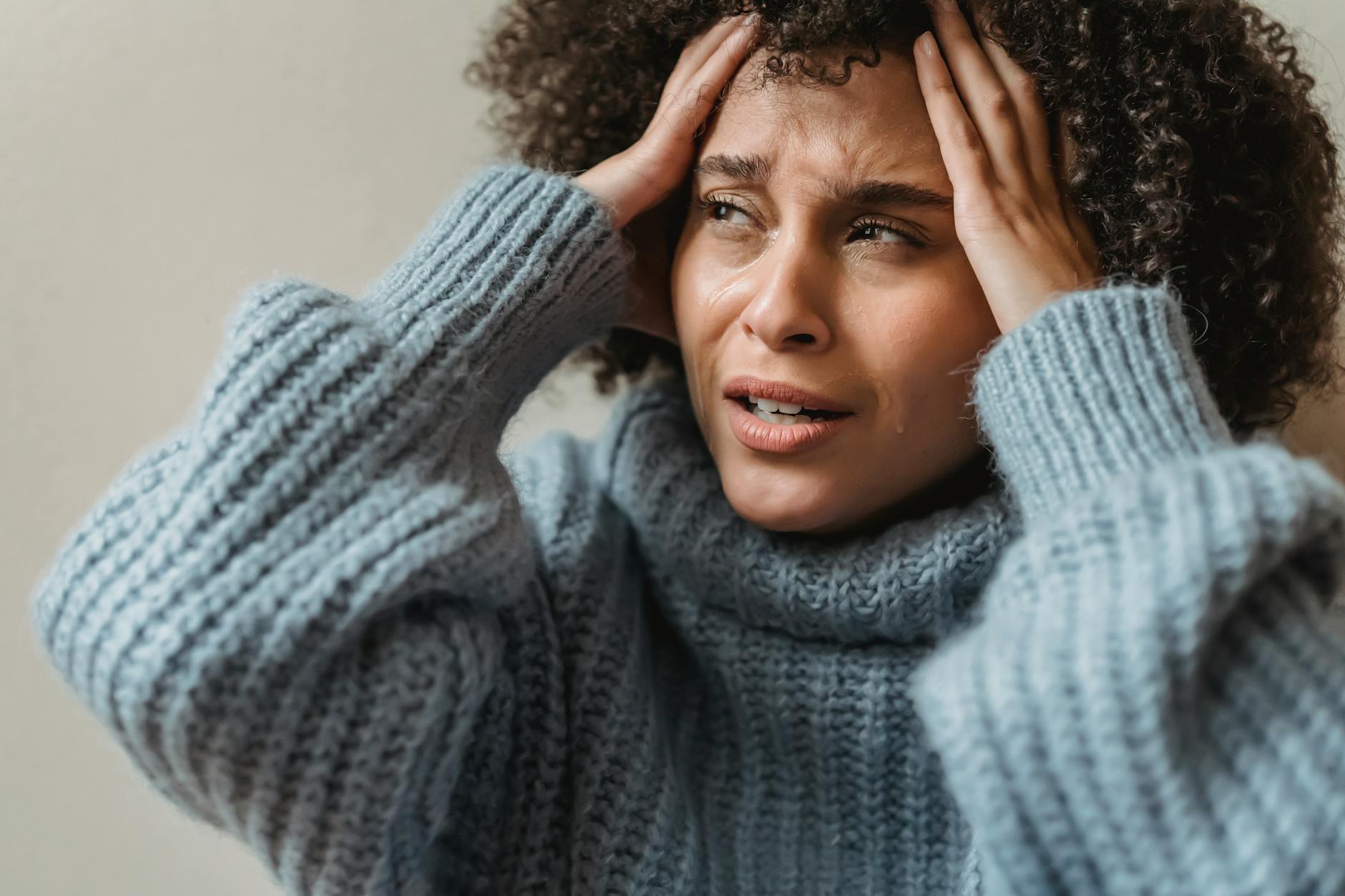 Worried African American female with brown curly hair crying and touching head against light wall