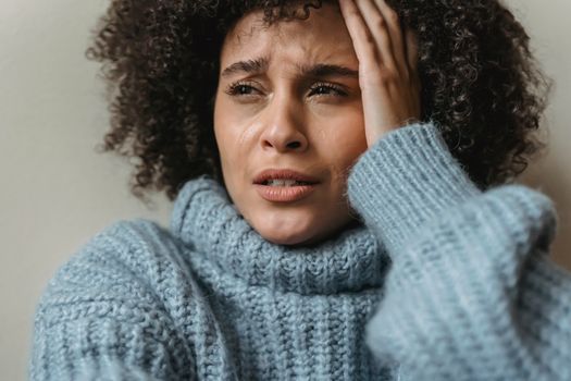 Unhappy African American woman with short curly hair in trendy warm sweater crying touching head and against light background