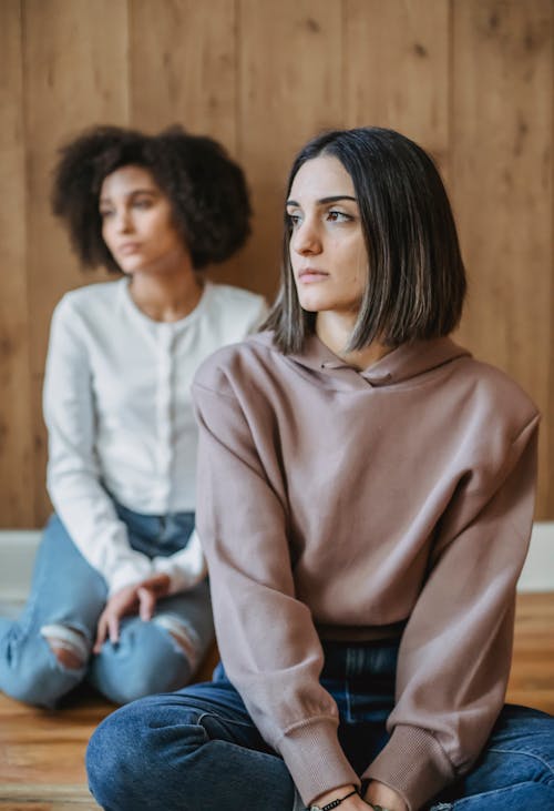 Thoughtful multiracial women in trendy clothes sitting on wooden floor and looking away in room in daytime