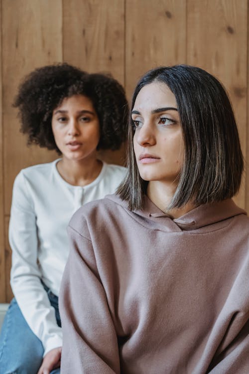 Pensive young multiethnic girlfriends in casual clothes sitting near wooden wall in light room