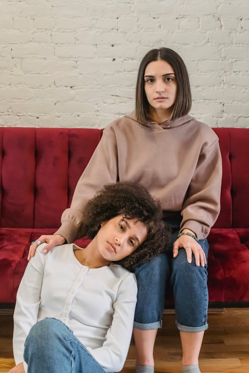 Young serious multiracial female friends sitting in light living room with lady laying head on laps of female on sofa near white brick wall and looking at camera