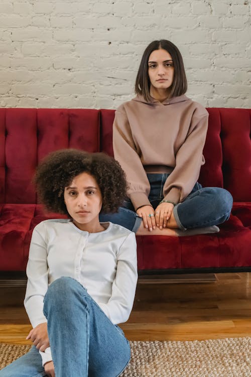 Young serious multiethnic female friends sitting in bright room with sofa near white brick wall and looking at camera