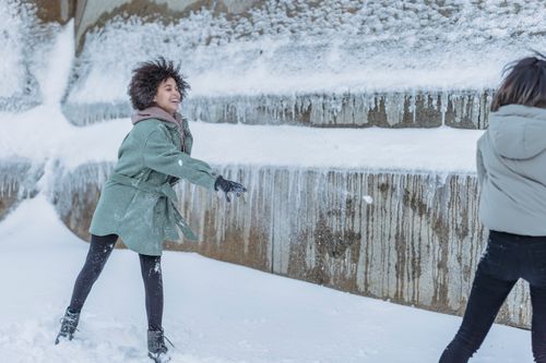 Free Smiling young multiethnic female friends in warm outerwear playing snowballs on snowy pavement near stone construction in winter day in city street Stock Photo