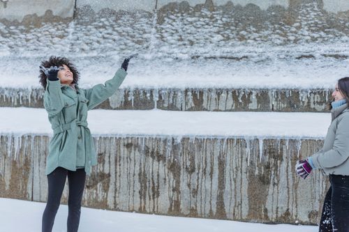 Free Happy young multiracial female friends in warm clothes playing snowballs on snowy walkway near building in city street in winter day Stock Photo