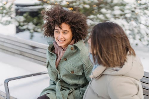 Free High angle of positive African American female with curly hair sitting on bench near girlfriend while spending time in winter park Stock Photo