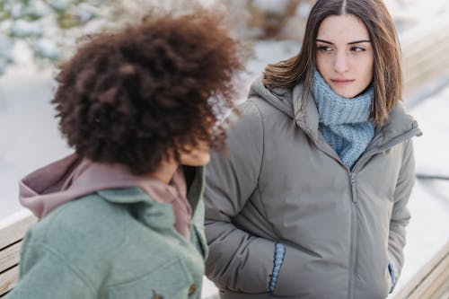 High angle of crop female listening to friend while sitting on wooden bench in winter park