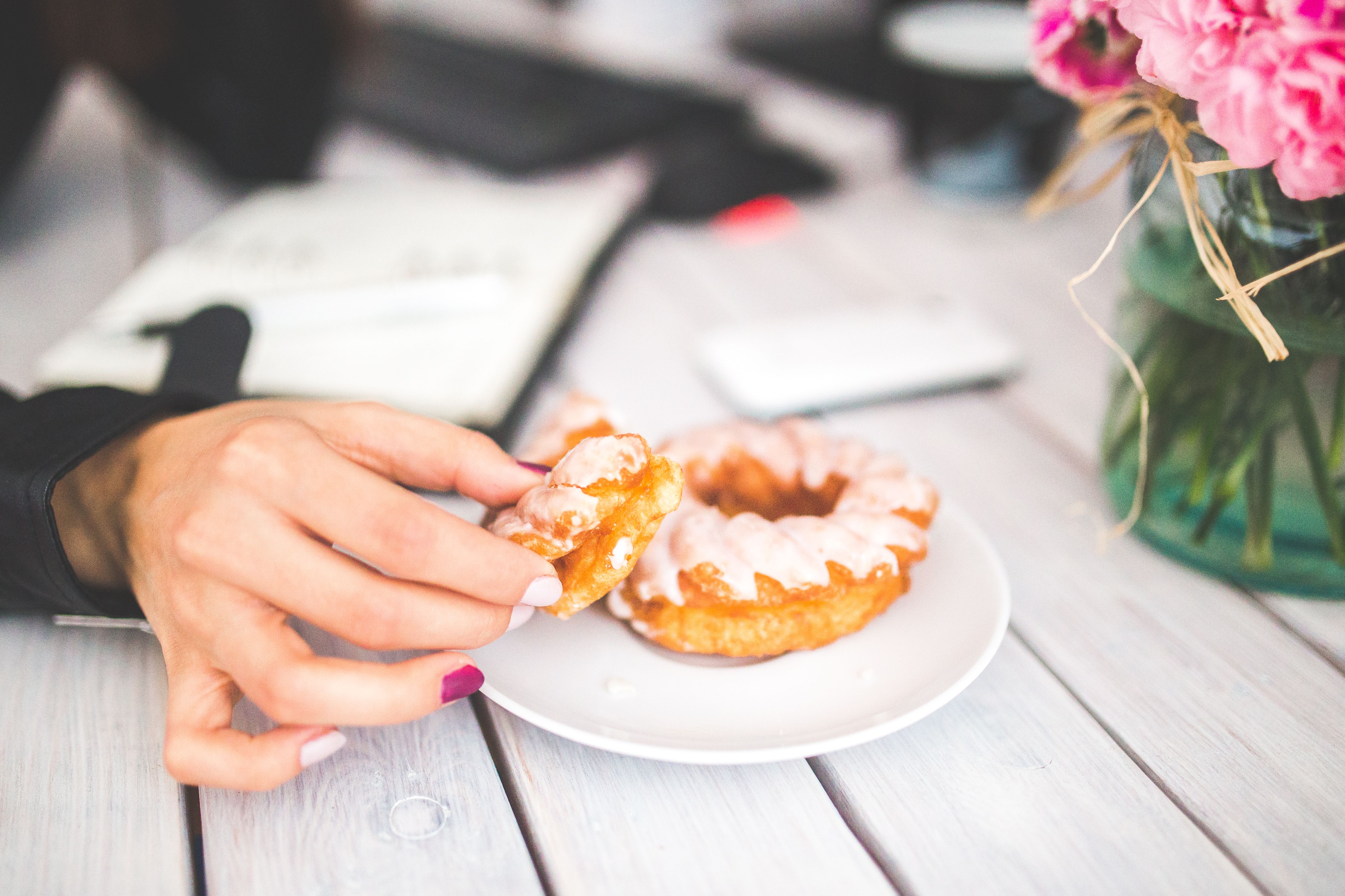 A woman's hand reaches for a glazed donut on a white plate indoors, showcasing a sweet treat moment.