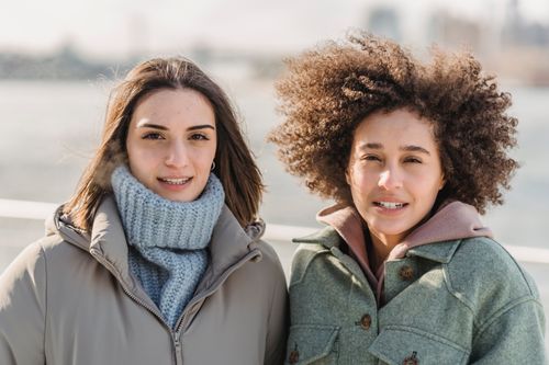Free Cheerful young multiethnic female friends in warm clothes standing together on city waterfront and looking at camera on sunny cold winter day Stock Photo