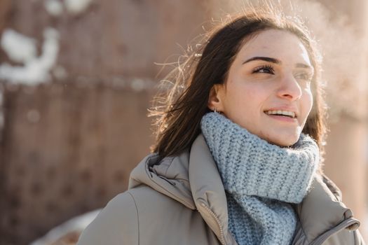 Content young brunette in blue sweater and warm jacket standing on snowy city street on cold sunny day and looking away