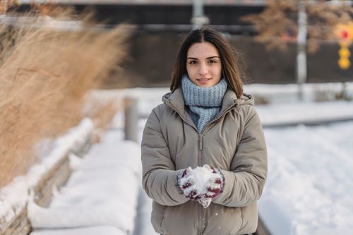 Free Content young female wearing warm clothes and mittens in warm clothes holding snow in hands spending winter day in city park and looking at camera Stock Photo