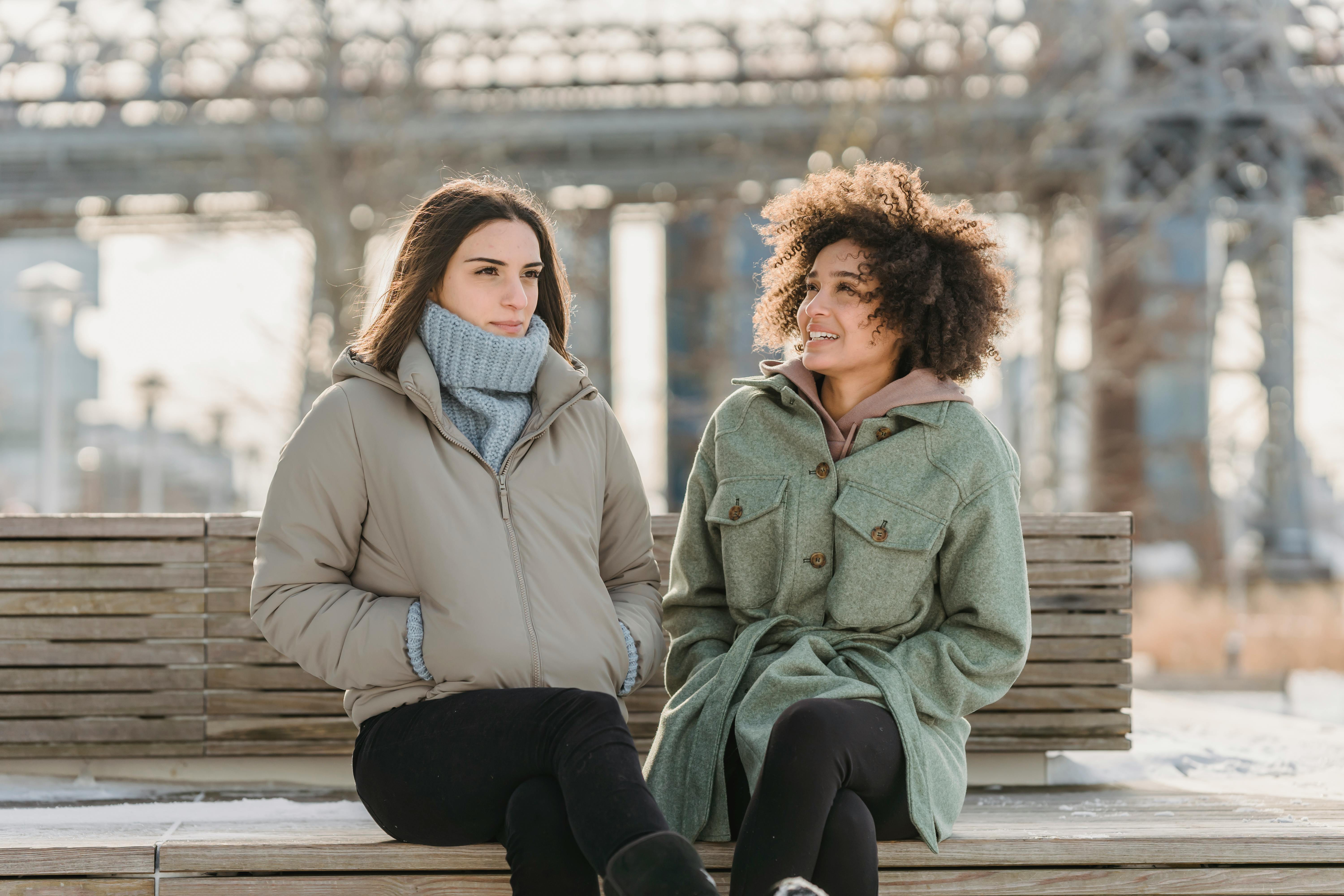 positive diverse women in outerwear sitting on bench on embankment