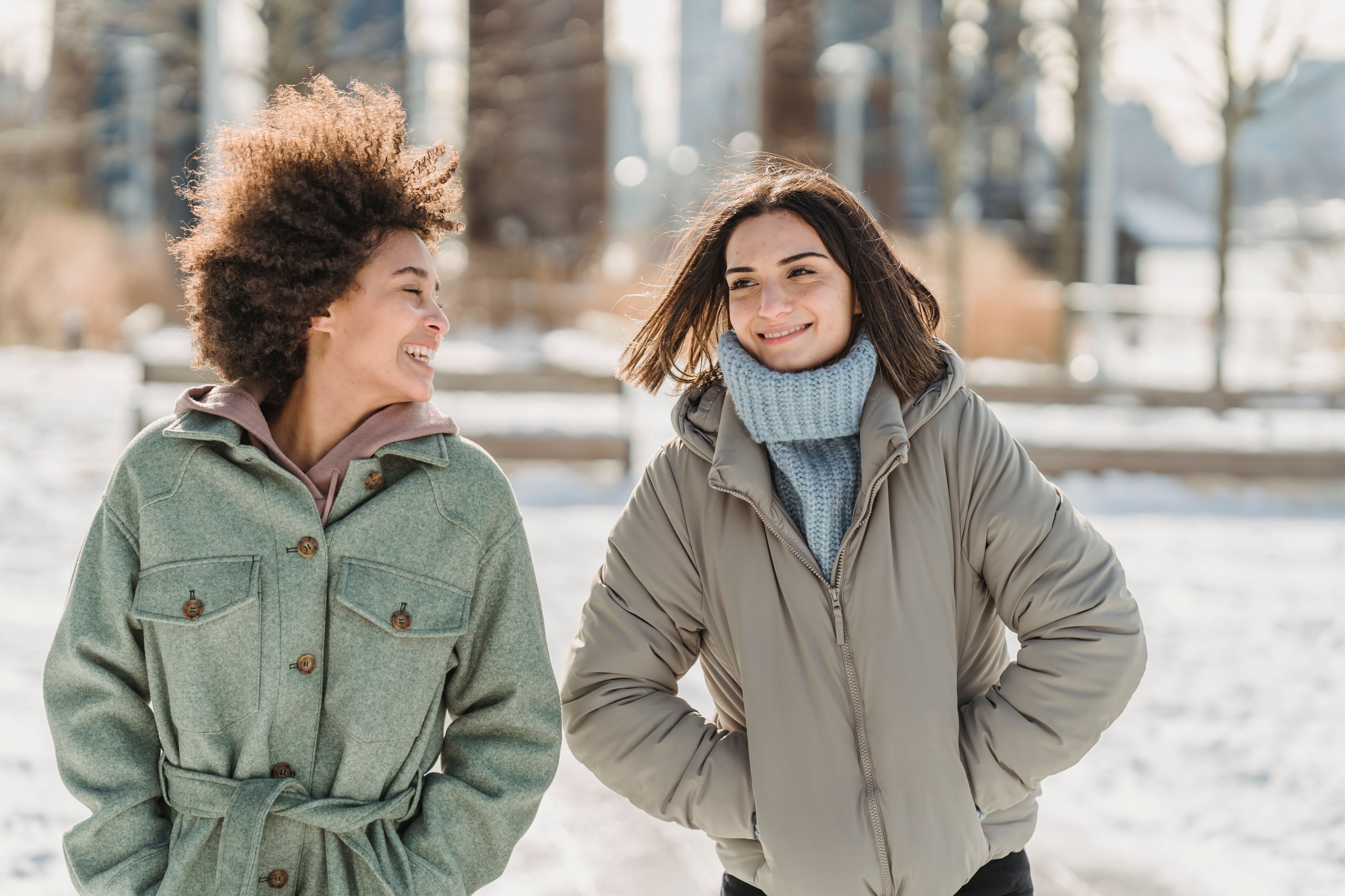 Carefree young multiethnic female friends in warm clothes walking together with hands in pockets on snowy city street and chatting