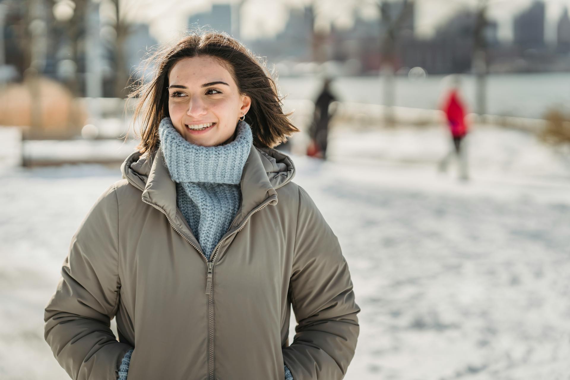 Content young brunette in blue knitted sweater and warm jacket standing with hands in pockets in snowy city park on cold winter day