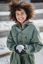 Smiling young ethnic female in warm coat and gloves holding snowball and looking at camera while spending sunny winter day in park