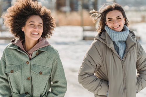 Carefree young multiracial female friends in warm wear walking together with hands in pockets on snowy city street on cold winter day
