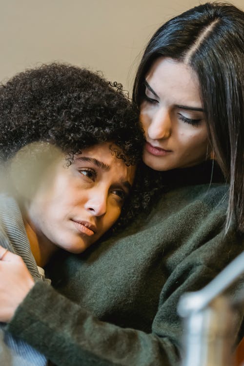 Young empathetic woman embracing and comforting upset ethnic female friend while sitting together at home