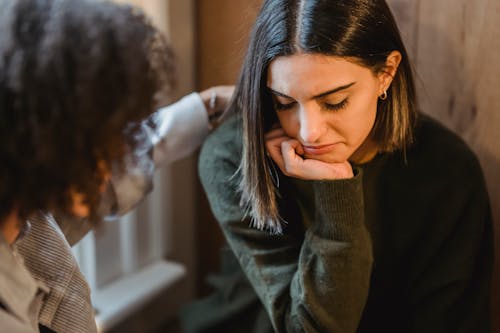 Crop woman tapping shoulder and comforting upset female friend while sitting at home together