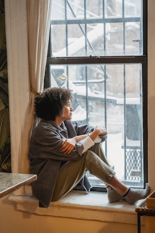 Sad young black woman sitting on windowsill and looking away pensively