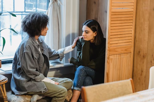 Calm young multiracial female best friends in casual clothes sitting near window and home and discussing discussing problems