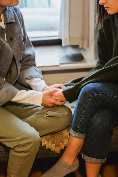 Side view of crop anonymous female best friends in casual clothes holding hands while sitting near window and discussing problems