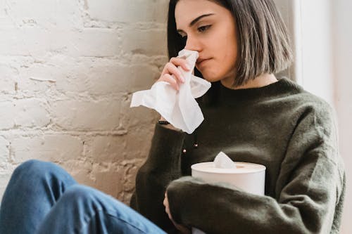 Crop young sad female with dark hair in sweater with tissue in hand crying and looking away sitting near white brick wall at home