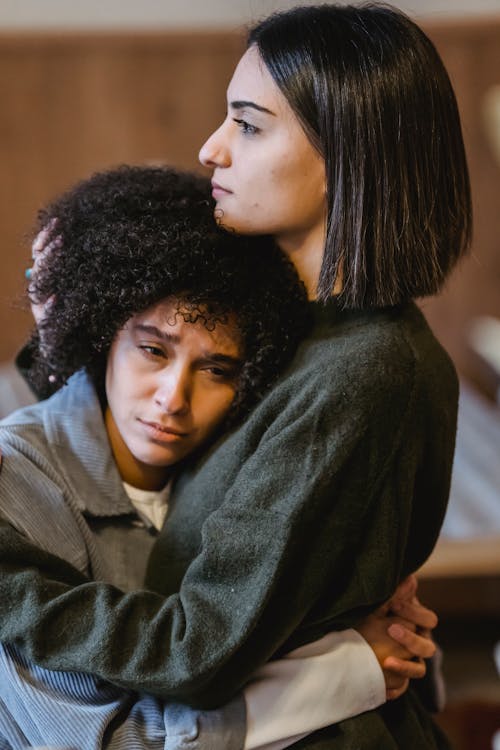 Side view of young lady with dark hair in casual clothes embracing and supporting sorrowful African American female friend at home