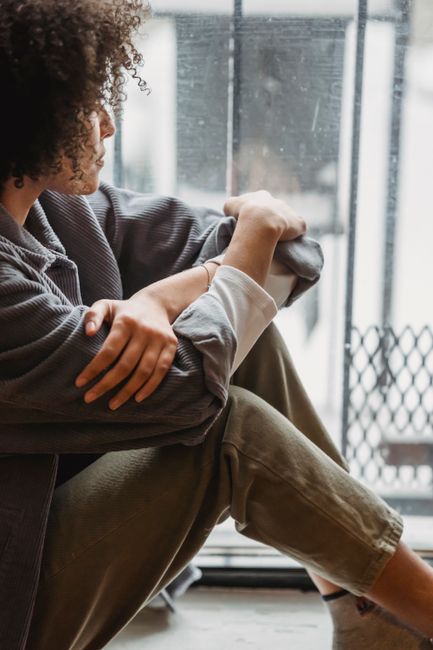 Side view of crop upset African American lady with curly hair in casual clothes sitting on windowsill and looking through window thoughtfully