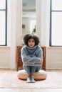Full body of sad young ethnic woman in casual outfit crying while sitting on pouf near windows and mirror near cabinet in light apartment and looking at camera