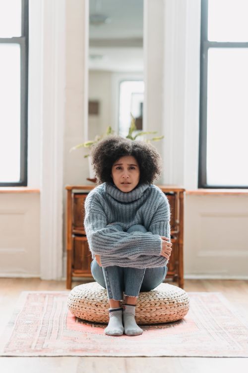 Free Full body of sad young ethnic woman in casual outfit crying while sitting on pouf near windows and mirror near cabinet in light apartment and looking at camera Stock Photo