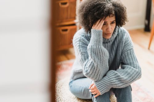 Free Young ethnic female in casual clothes sitting on pouf while crying with hand on head in light flat Stock Photo