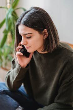 Serious young woman in casual outfit sitting in light room while talking on cellphone near green plants