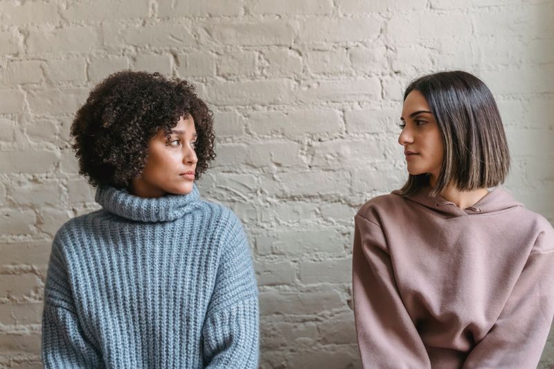 Emotionless young multiethnic women in casual outfit looking at each other near white brick wall in bright house