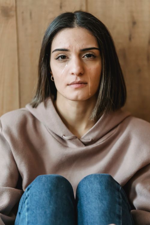 Free Young woman in casual clothes sitting near wooden wall in light apartment and looking at camera while crying Stock Photo