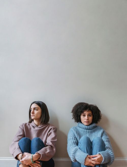 Free Pensive young multiethnic female friends in casual clothes sitting together while leaning on white wall in light apartment Stock Photo