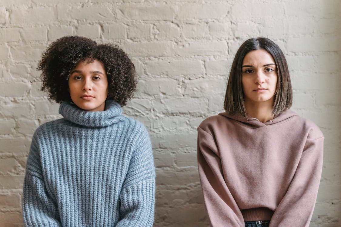 Diverse female friends in warm clothes looking at camera while sitting near white colored brick wall in light room
