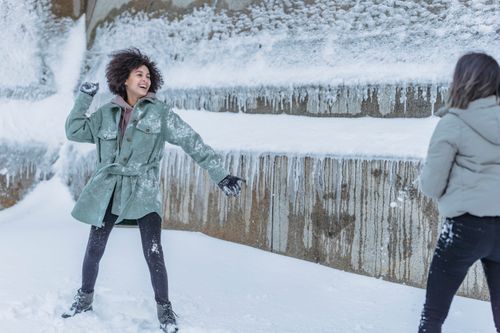 Free Laughing female friends in warm clothes standing near stone wall covered with snow and playing snowballs in winter Stock Photo