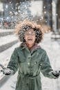 Young ethnic female in warm clothes with curly hair laughing while tossing up snow on blurred background in city