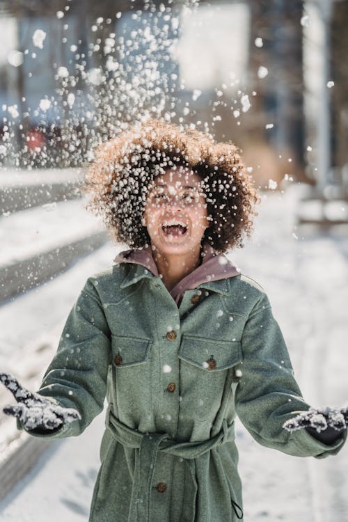 Free Young ethnic female in warm clothes with curly hair laughing while tossing up snow on blurred background in city Stock Photo
