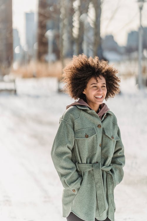 Cheerful ethnic female in outerwear looking away while standing on snowy sidewalk with hands in pockets on street in winter day on blurred background