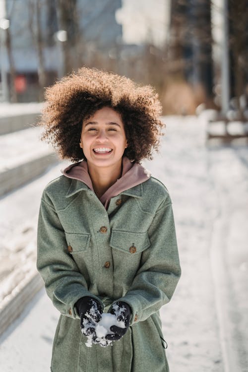 Positive ethnic female with snowball in hands looking at camera while playing on snowy sidewalk on blurred background in winter day