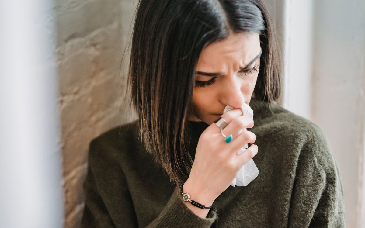 Despaired female with dark hair in casual clothes standing near wall with tissue in hand in light room at home