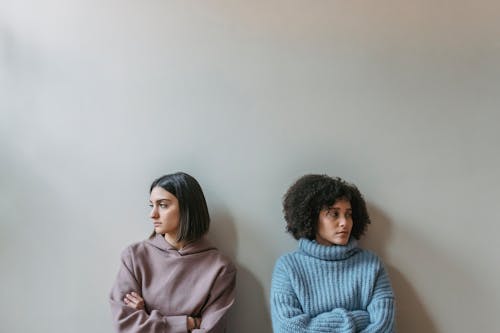 Sad multiracial female friends with crossed arms looking away and standing near white wall while having conflict in light room at home